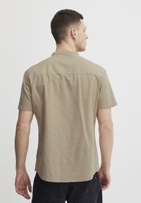 Kortermet beige skjorte med krage, teksturert stoff og rett kant, vist fra baksiden på en person.