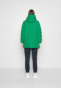 Veste matelassée verte avec capuche, épaules tombantes et poignets élastiques, associée à un pantalon de jogging bleu marine et des sneakers blanches.