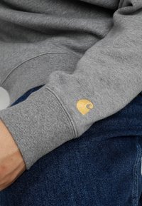 Šedá mikina s žebrovaným manžetovým lemem a malým zlatým logem. Textura vypadá jemně, doplněná tmavými džíny.