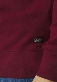 Sudadera burdeos con una textura suave, que presenta una pequeña etiqueta negra cerca del dobladillo y puños y cintura de canalé.