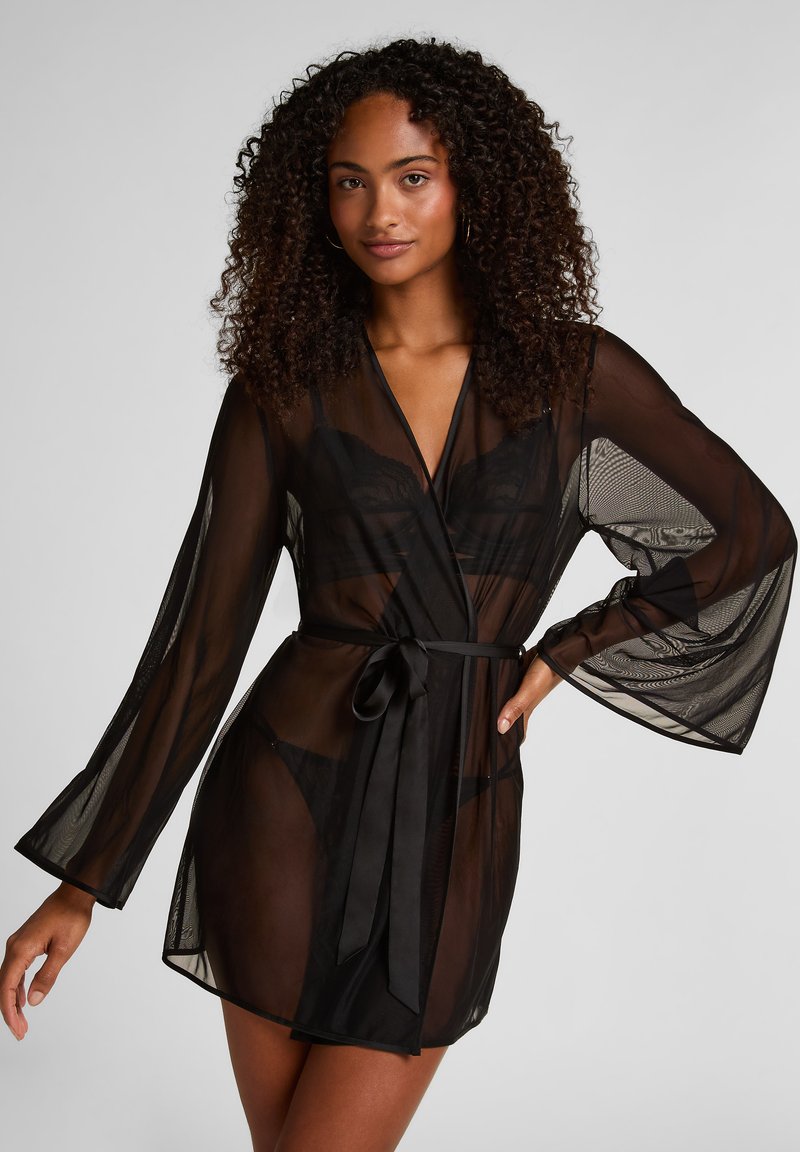 Γυναίκα με σγουρά μαλλιά που φοράει διαφανή μαύρη ρόμπα με ζώνη στη μέση πάνω από μαύρα εσώρουχα, ποζάροντας με το ένα χέρι στη μέση μπροστά σε λευκό φόντο.