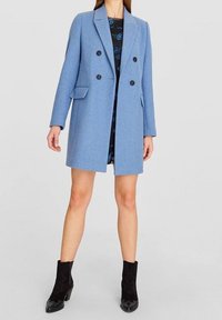 Dubbelknäppt blå kappa i texturerad tyg, med tre framfickor, spetsig slagkrage och svarta knappar. Bärs över en mönstrad klänning.