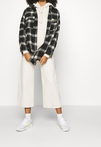 Chemise en flanelle à carreaux noirs et jaunes portée par-dessus un sweat à capuche beige clair, associée à un pantalon large ivoire et des baskets blanches avec des accents néon.