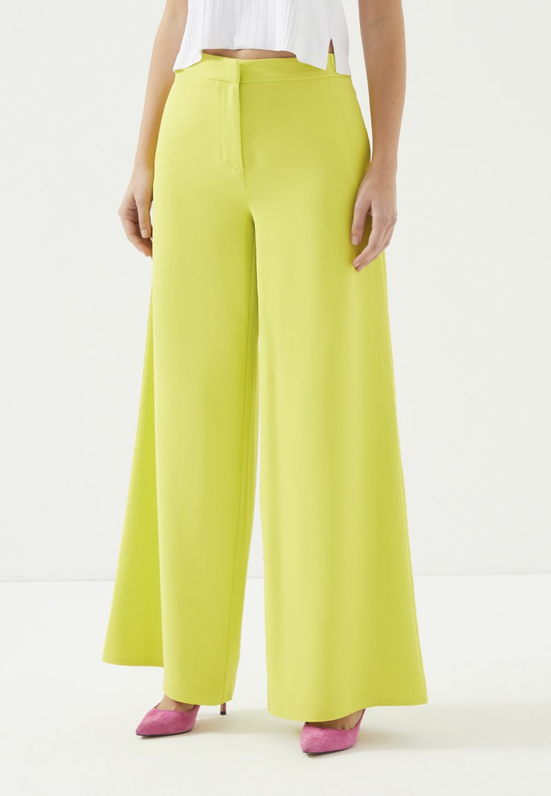 Wijde broek in fel limoen groen polyester, met een hoge taille, een gladde textuur en een verborgen rits aan de achterkant. Gecombineerd met roze hakschoenen.