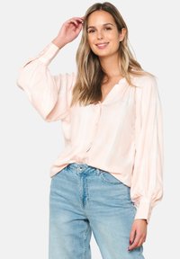 Light pink blouse with a V-neck, button closure, and long, puffed sleeves. Features a subtle check pattern, worn with light blue jeans.