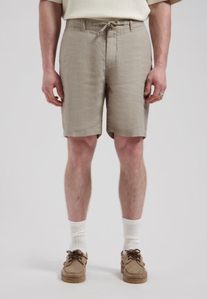 Man draagt beige shorts met trekkoord, witte sportsokken, bruine mocassinschoenen, staand tegen een effen lichte achtergrond.