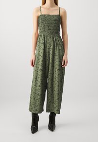 Khaki zelený overal s nařaseným živůtkem a tenkými ramínky, s květinovým vzorem, širokými nohavicemi a stylizovaný s černými špičatými kotníkovými botami.