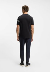 Schwarzes kurzärmliges Hemd mit einem weißen Akzentstreifen auf der linken Schulter, kombiniert mit marineblauen Hosen und schwarzen Sneakers.