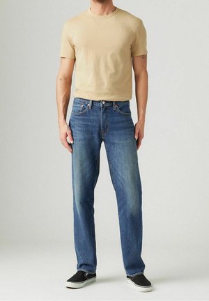 Mand iført beige T-shirt med korte ærmer, blå jeans med lige ben og sorte slip-on sko står foran en ensfarvet grå baggrund.