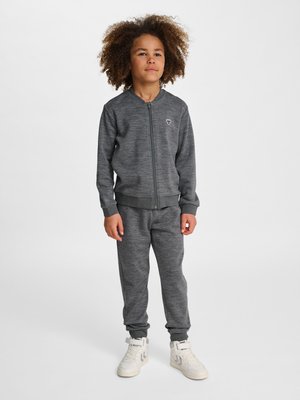 Barn med krøllet hår iført grå lynlås-sweater, matchende bukser og hvide sneakers, der står mod en ensfarvet lys baggrund.