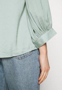 Helles grünes Satin-Bluse mit Puffärmeln, Raffungen an den Manschetten, kombiniert mit verblassten blauen Jeans, die einen lässigen Look präsentieren.