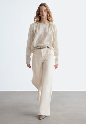 Ung kvinde iført beige stribet bluse, bukser med vidde og høj talje samt spidse hæle, gående foran en ensfarvet hvid baggrund.