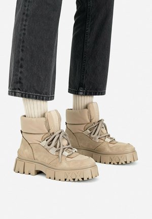 Beige grove veterschoenen gedragen met crèmekleurige geribbelde sokken en zwarte cropped broek op een witte achtergrond.