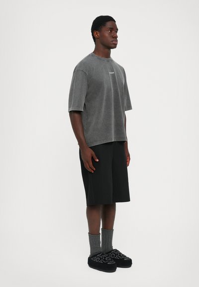 Man staat gekleed in een gewassen grijze oversized T-shirt, knielange zwarte shorts, grijze sokken en zwarte instapschoenen met een patroon.