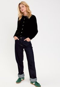 Cardigan nero a bottoni, jeans in denim scuro con risvolti, e sneakers verdi con dettagli bianchi, che creano un look casual e sovrapposto.