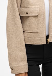 Giacca di lana beige con design cropped, dotata di due grandi tasche frontali, hardware dorato e chiusura a zip. Morbido al tatto e vestibilità sagomata.
