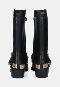 Botas de cuero negro hasta el tobillo con un caña alta, acentos de hardware metálico, cremalleras laterales y tacones apilados. Las correas decorativas con hebilla mejoran el diseño.