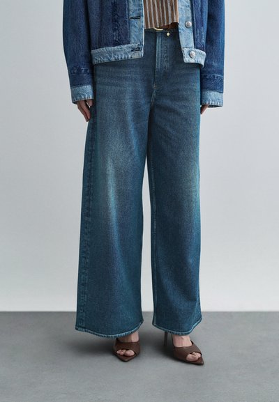 Jean à jambes larges en denim bleu délavé avec une taille haute, présentant des coutures visibles et une coupe décontractée. Associé à des talons marron.