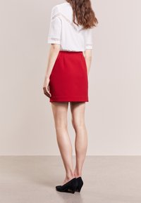 Falda roja ajustada con una textura suave, combinada con una camisa blanca que presenta detalles de encaje en los hombros. Tacones de ante negros completan el look.