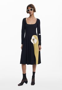 Schwarzes langärmeliges Kleid mit tailliertem Oberteil und ausgestelltem Rock, das auf der linken Seite eine große gelbe und weiße Grafik aufweist. Glatte Textur.