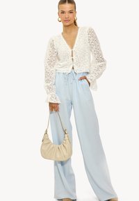 Top en dentelle blanche avec un décolleté en V profond et un nœud à l'avant, associé à un pantalon large bleu clair. Le modèle porte un sac à épaule beige doux.