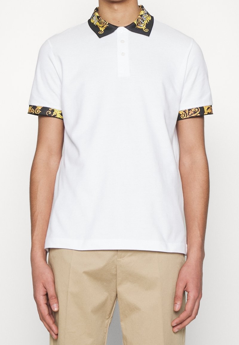 Homme portant un polo blanc à manches courtes avec un col et des bords de manches ornés de motifs noirs et dorés, associé à un pantalon beige.