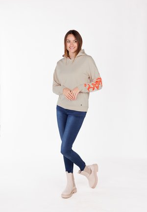 Haut à capuche beige avec une poche avant et un graphique orange sur la manche droite. Associé à un jean skinny bleu foncé et des bottines beiges.