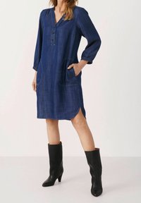 Vestido de mezclilla con una textura suave, escote en V, mangas tres cuartos, dos bolsillos delanteros, aberturas laterales y un corte ligeramente holgado.