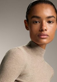 Kvinde med glat hud og trukket tilbage hår iført en beige ribbet turtleneck sweater, der ser direkte fremad mod en neutral baggrund.