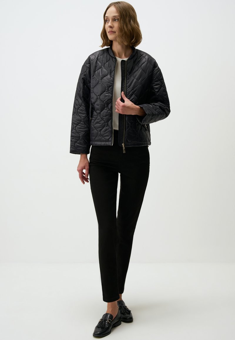 Veste bomber noire matelassée avec fermeture éclair à l'avant, présentant un col rond et des manches larges, associée à un pantalon noir et des mocassins.