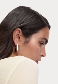 Gouden druppelvormige oorbellen met een glanzend, glad oppervlak. Heeft een geometrisch ontwerp en is dicht bij het oorlel gemonteerd, waardoor hun vorm wordt benadrukt.