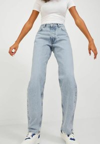 Šviesiai mėlynos aukštos juosmens džinsai su tiesiomis kojomis, pasižymintys išblukusiu tekstūru ir kontrastingomis siūlėmis, derinami su baltais sportbačiais.