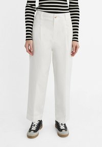 Pantalon blanc à jambes larges en tissu léger avec des plis marqués, porté avec un haut rayé noir et blanc et des baskets noires.