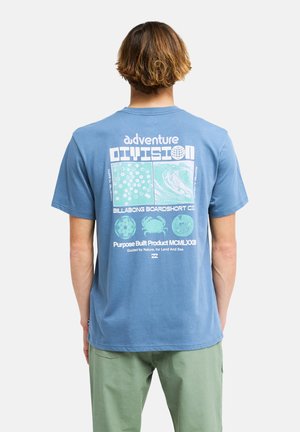 Person mit kurzen braunen Haaren trägt ein blaues T-Shirt mit natur- und ozeanbezogenen Illustrationen und Text auf dem Rücken.