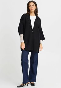 Black oversized blazer with notch lapels and a single button. Underneath, a white top with lace sleeves. Paired with wide-leg dark jeans and black shoes.