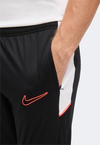 Ръка в джоба с черен панталон Nike с червен логотип swoosh и бял и червен страничен панел.