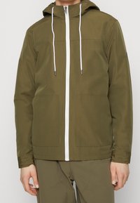 Chaqueta con capucha color caqui, con cremallera frontal blanca y cordones, que cuenta con dos bolsillos frontales y un acabado suave y mate.