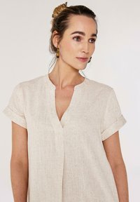 Chemise courte à manches courtes beige clair avec un col en V, en tissu texturé, et des manches à revers, présentant de subtils éclats tout au long.