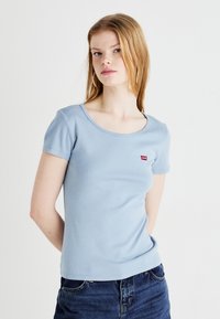 Világoskék, testhezálló póló rövid ujjakkal, kerek nyakkivágással és piros Levi's logóval a bal mellrészen, kék farmernadrággal párosítva.