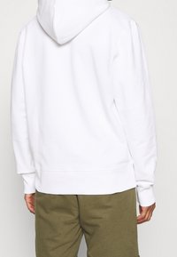 Felpa bianca con cappuccio e tasca a marsupio, polsini e orlo a costine, abbinata a pantaloni corti verde oliva. Texture morbida e design casual.