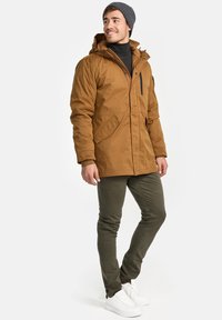Brun isolert parkas med glidelås og trykknappelukking, store lommer og avtagbar hette, kombinert med olivengrønne bukser og hvite joggesko.