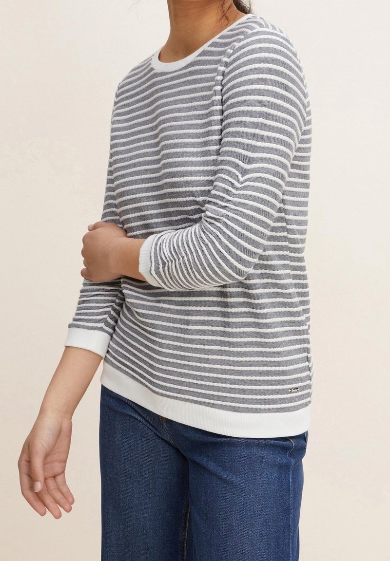 Grå och vit randig tröja med rund halsringning och ribbade ärmmuddar, matchad med blå jeans. Tyget verkar något texturerat.