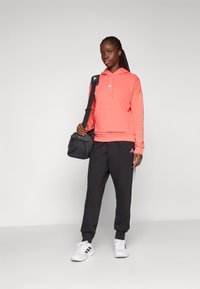 Korallfärgad hoodie med en känguruficka, parat med svarta sweatpants med vita ränder. Svart axelväska, vita sneakers med svarta detaljer.