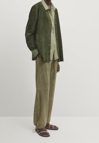Personne portant une chemise verte à boutons superposée et une veste, avec un pantalon ample olive et des sandales marron, debout devant un fond uni.