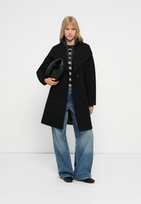 Cappotto di lana nera con un ampio colletto strutturato, indossato su una maglietta a righe, abbinato a jeans a zampa d'elefante e una borsa nera arrotondata.