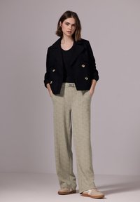Chaqueta negra de corte cruzado con botones dorados, combinada con pantalones de pierna ancha de patrón verde claro y zapatillas beige con detalles en blanco.