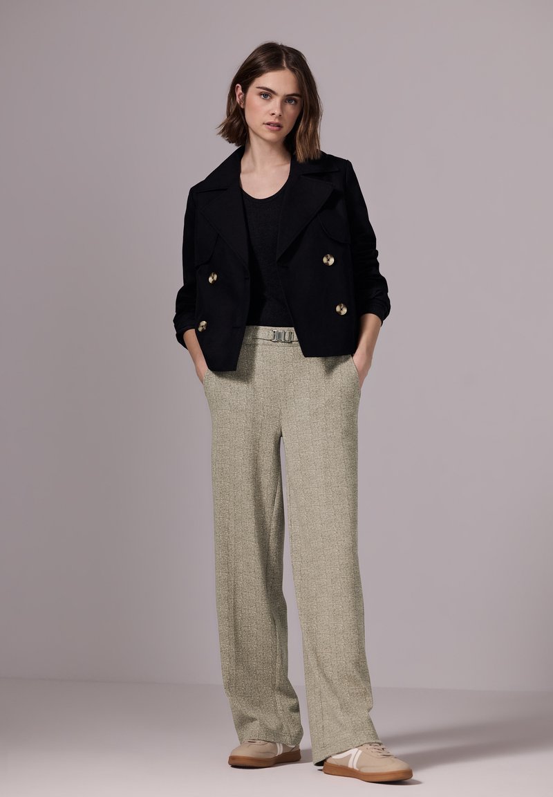 Chaqueta negra de corte cruzado con botones dorados, combinada con pantalones de pierna ancha de patrón verde claro y zapatillas beige con detalles en blanco.