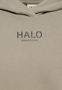Beige hoodie gemaakt van zachte stof met een voorzak in kangoeroestijl, een capuchon met trekkoord en donkergeprinte tekst: "HALO 196819721981."