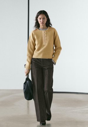 Maglione a costine color senape con scollo abbottonato, abbinato a pantaloni marroni a vita alta; tiene una borsa nera testurizzata; design semplice.