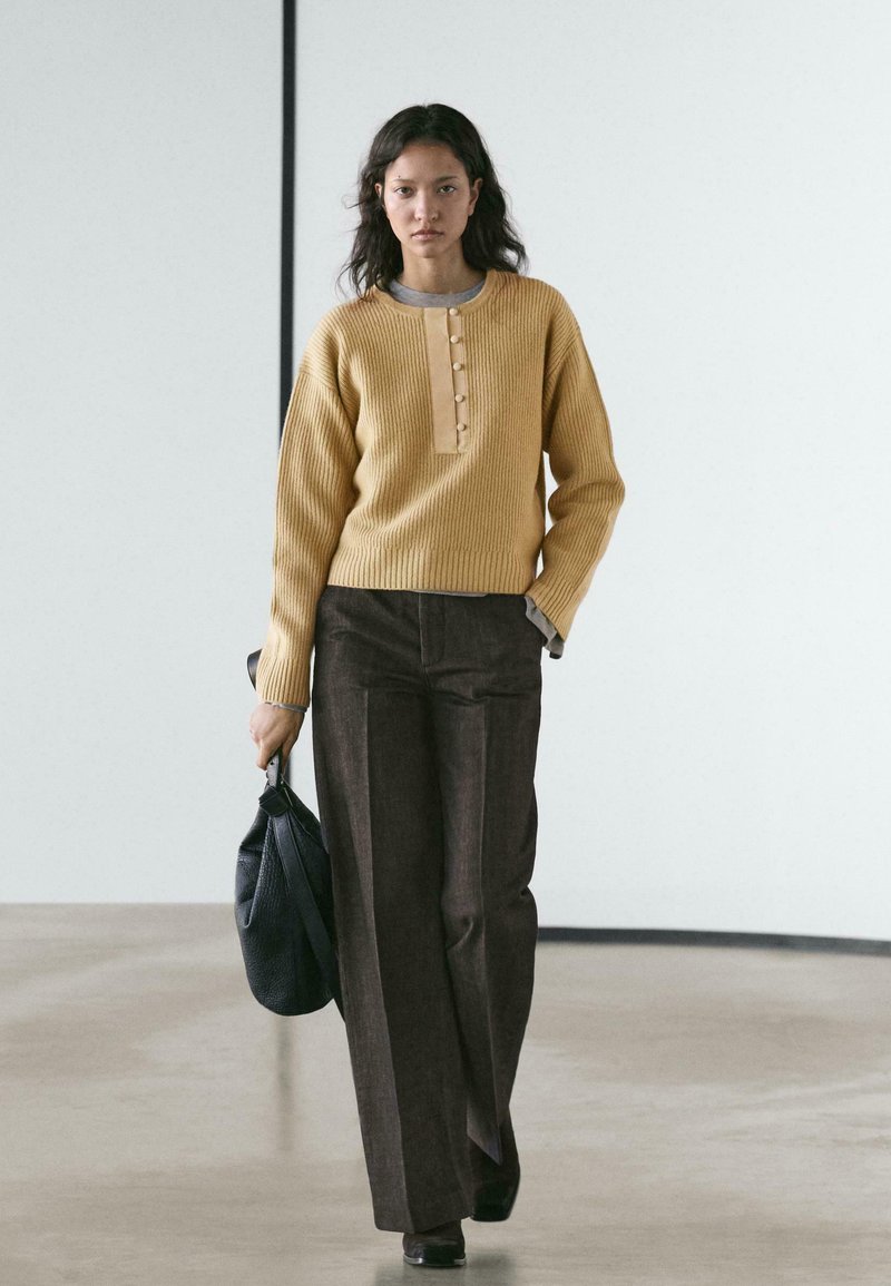 Maglione a costine color senape con scollo abbottonato, abbinato a pantaloni marroni a vita alta; tiene una borsa nera testurizzata; design semplice.